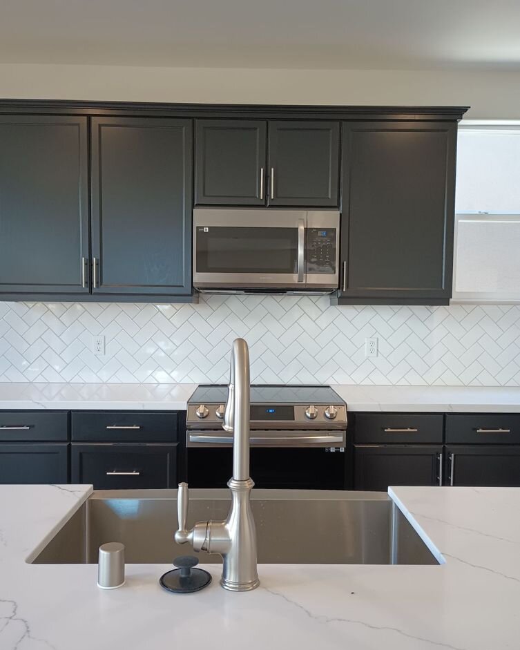 Charcoal shaker kitchen cabinets with a built-in microwave and herringbone tile backsplash, seen across a quartz island sink