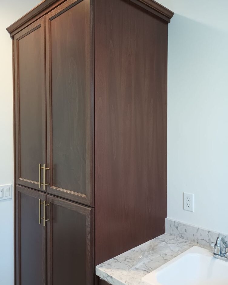 Tall dark wood linen cabinet with crown molding and brass bar pulls beside a bathroom vanity top